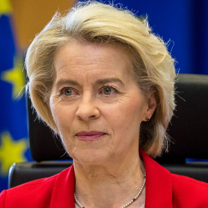 Europa abierta - Primer plano de Ursula von der Leyen, presidenta de la Comisión Europea, con chaqueta roja, collar de perlas y la bandera de la UE al fondo.