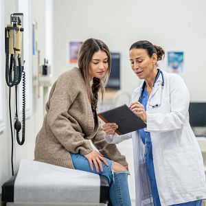 Marca España - Médica con bata blanca y estetoscopio muestra una tableta a paciente en camilla, en consulta médica iluminada. Se observa tensiómetro y escritorio con monitor.