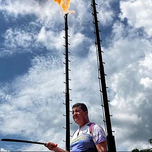 Hora América - Hombre con camiseta deportiva sostiene un palo, frente a dos altas estructuras industriales, una de ellas en llamas.