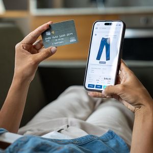 Economía de bolsillo - Persona de piel oscura comprando online con tarjeta de crédito y móvil. Se ve una app de ecommerce con jeans y precio. Hora: 14:23.