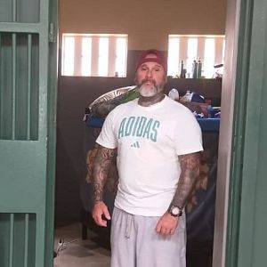 Hora América - Plano medio de un recluso con brazos tatuados, camiseta blanca y gorra, de pie junto a una puerta de celda metálica numerada como CELL C2.