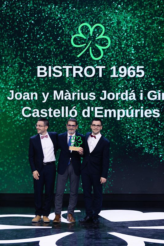Ceremonia de premiación: tres hombres en traje, uno con un premio con forma de trébol, frente a una pantalla verde que menciona al restaurante 'BISTROT 1965'.