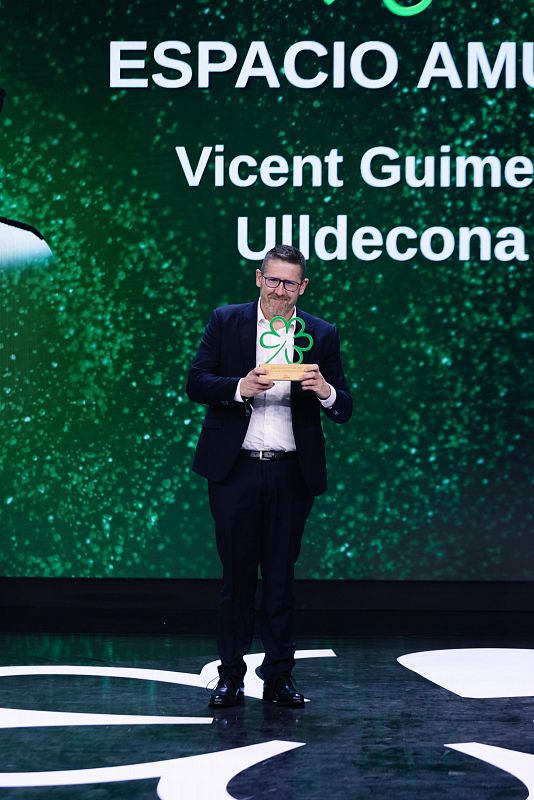 Vicente Guime de Ulldecona recibe una estrella verde por prácticas sostenibles en un escenario con pantalla verde que muestra su nombre y "ESPACIO AMU".