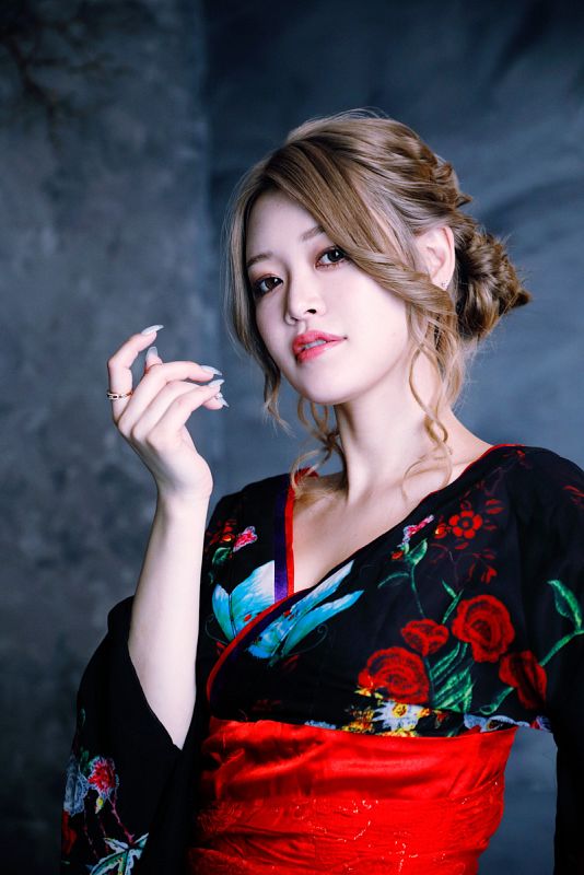 Retrato de mujer con kimono negro y rojo con estampado floral, cabello rubio recogido en moño bajo y expresión seria. Uñas largas y anillo en un dedo.