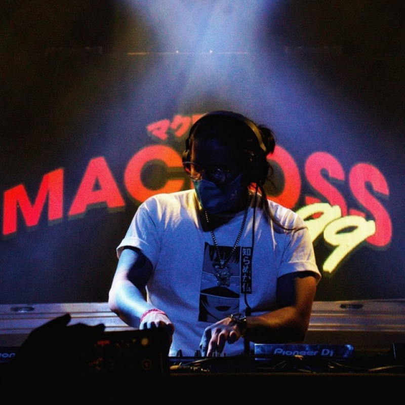 DJ con gafas y mascarilla actuando con una mesa de mezclas Pioneer DJ.  Pantalla con "MACROSS 82-99" al fondo. Lleva camiseta con estampado anime y accesorios.