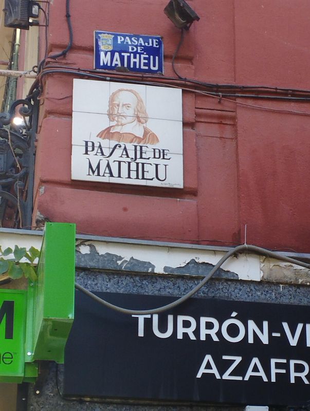 Letreros cerámicos con el nombre del pasaje y retrato; cartel de negocio de turrón y azafrán parcialmente visible; cables y cámara de seguridad en pared deteriorada.