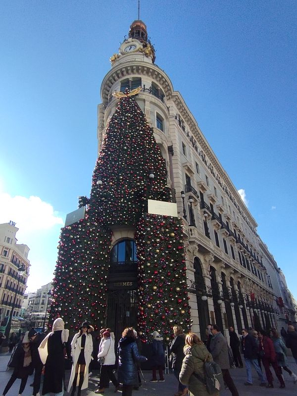 Imponente árbol navideño con adornos rojos, dorados y plateados en esquina de edificio clásico con fachada clara, balcones y reloj. Se lee "HERMES" en la entrada.