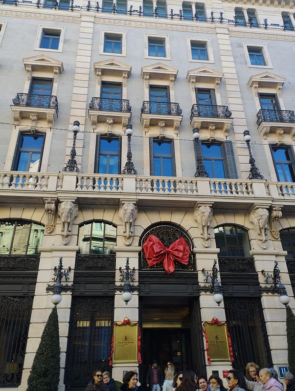 Decoración navideña en la fachada de un edificio con balcones, farolas y dos cabezas de elefante.  Un grupo de personas se encuentra en la entrada, adornada con un gran lazo rojo.