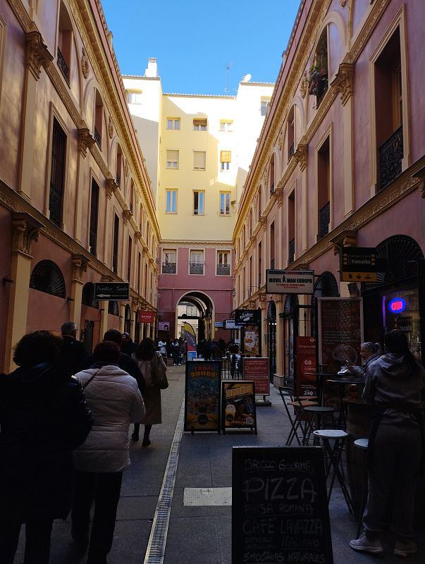 Pasaje comercial con edificios de varios pisos, personas caminando, y letreros de negocios como 'Chacaito' y 'Movil & Man Europa', ofreciendo pizza, café y smoothies.