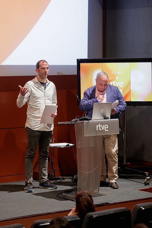 Dos hombres en una gala de bienvenida, uno hablando y otro en un atril con un portátil, en un espacio con el logo de RTVE. Se ve parte del público.