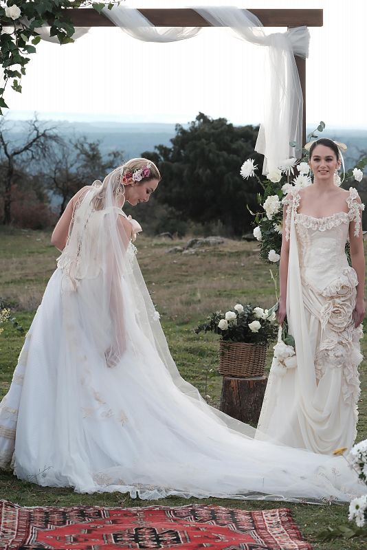 Presentación de dos vestidos de novia: uno blanco bordado con cola, otro crema con volantes.  Ambas modelos posan bajo un arco floral sobre alfombra roja.