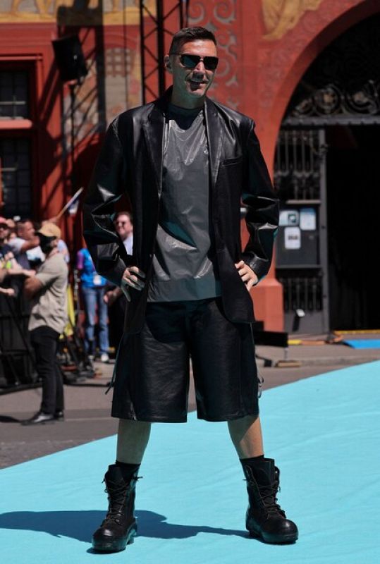 Persona con chaqueta negra, camiseta gris y pantalones cortos, posando en alfombra turquesa.  Contexto sugiere evento similar a alfombra roja.