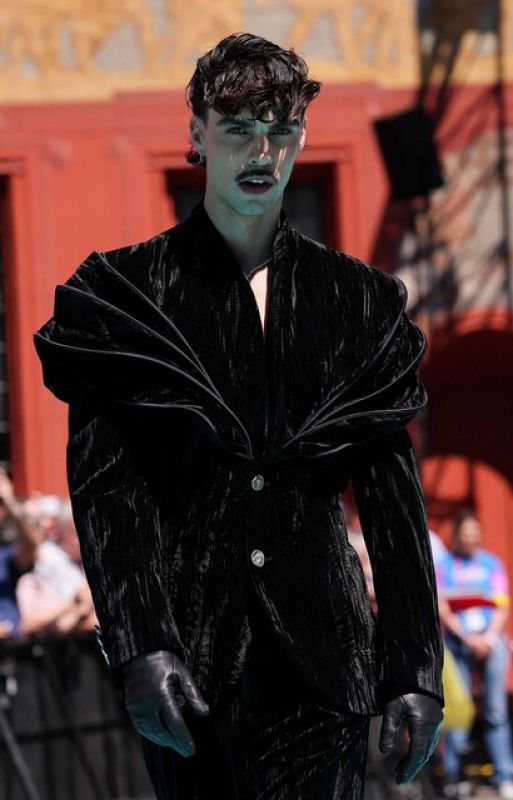 Persona con traje de terciopelo negro, guantes y pendientes, en una alfombra turquesa durante un evento al aire libre.