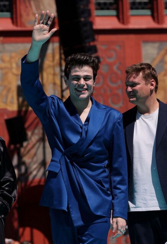 Blanca Paloma en la Alfombra Turquesa con un traje azul cruzado, anudado a la cintura y varios anillos. Saluda con la mano derecha; al fondo, una estructura rojiza y parte de otra persona.