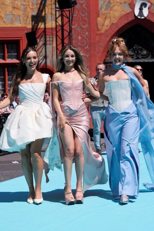Tres mujeres con vestidos de diferentes colores (blanco, rosa pálido y azul) posan sobre una alfombra turquesa.  Se aprecia un edificio decorado con murales al fondo.