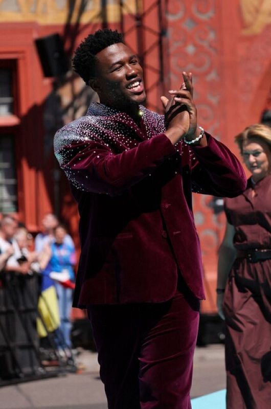 Hombre en traje granate con pedrería aplaude; tras él, mujer con vestido marrón; fondo con público y alfombra turquesa.