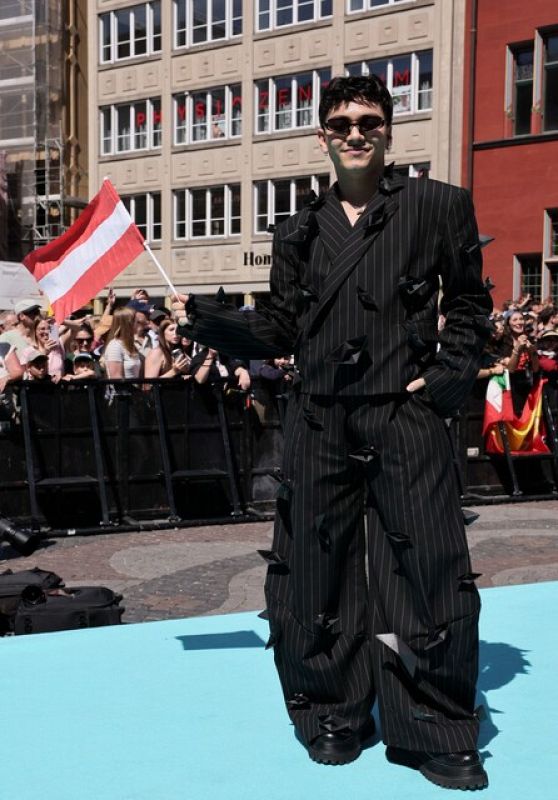 Hombre con traje de rayas negras y figuras geométricas, sosteniendo una pequeña bandera, posa en una alfombra turquesa.  Zapatos de plataforma.