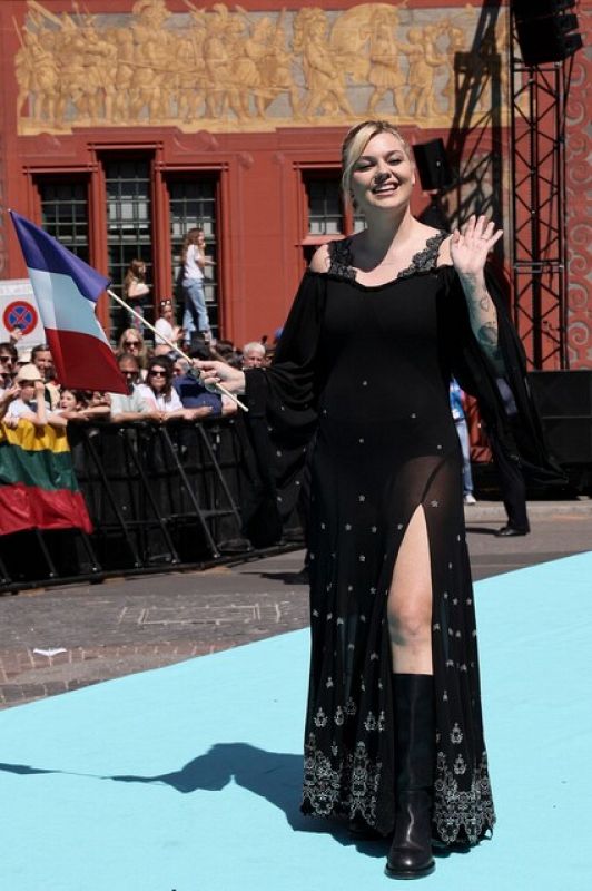 Mujer en vestido negro y botas altas, con bandera francesa, camina por alfombra turquesa en evento con otras banderas y personas.