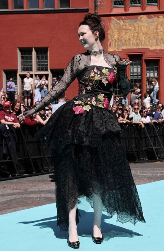 Persona con vestido negro gótico, bordado floral rosa, mangas transparentes y falda asimétrica, posa en alfombra turquesa con zapatos negros de tacón bajo y moño alto.