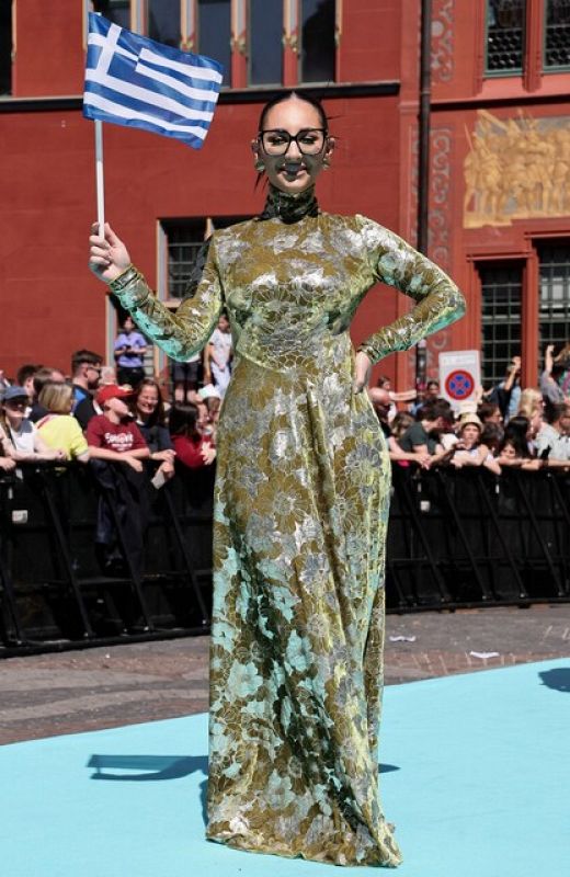 Mujer con vestido largo floral, gafas oscuras y bandera griega, sonríe en alfombra turquesa. Fondo con edificio de ladrillo.