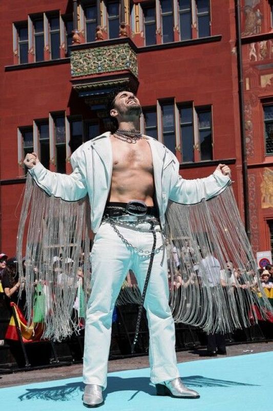 Representante de Armenia en Eurovisión 2025 posa en alfombra turquesa con chaqueta blanca, pantalones blancos y botas plateadas; pose dramática, fondo con público y banderas.