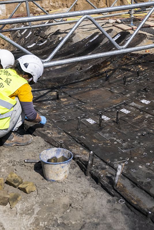Arqueólogo examina tablones de madera numerados durante excavación.  Herramientas y cubo presentes.  Estructura metálica protege el sitio.