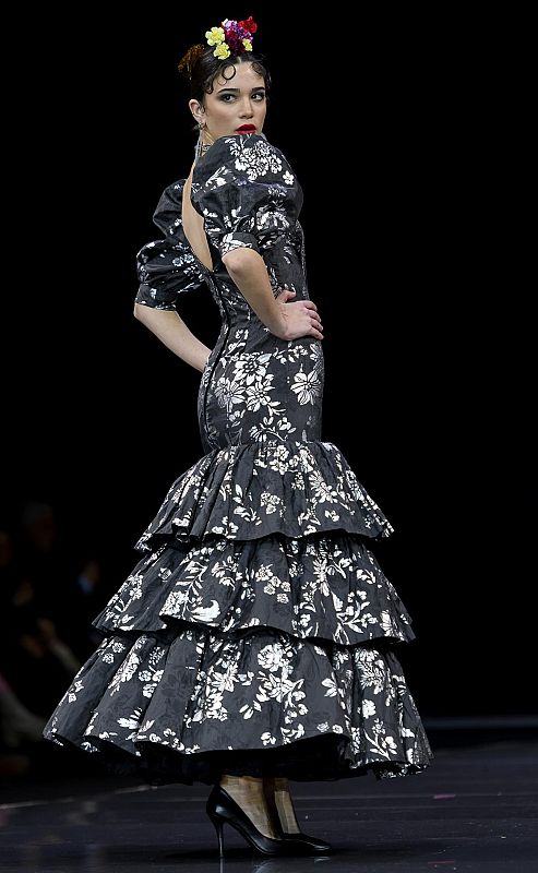 Vestido de flamenca contemporáneo gris y plateado con estampado floral, mangas abullonadas y falda con volantes. Modelo con recogido y zapatos negros desfilando en pasarela.