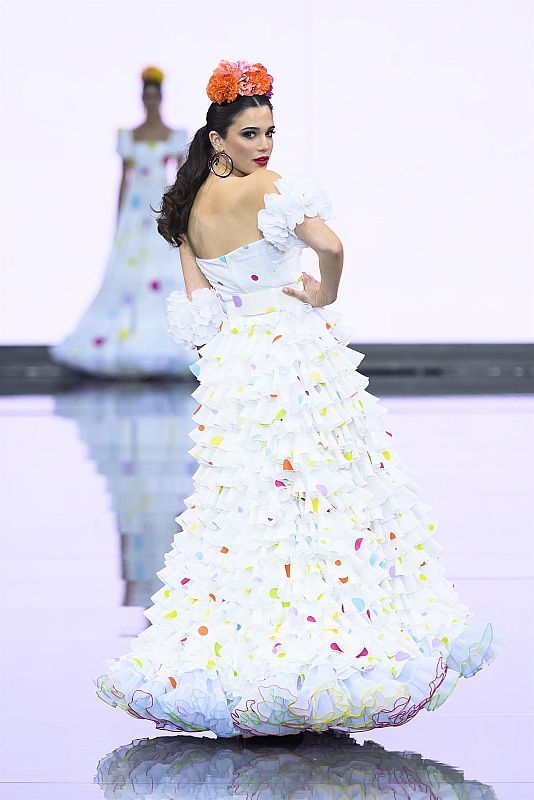 Vestido blanco con lunares, múltiples volantes y escote off-the-shoulder. Modelo con pose dinámica en pasarela brillante; reflejo del traje en el suelo.