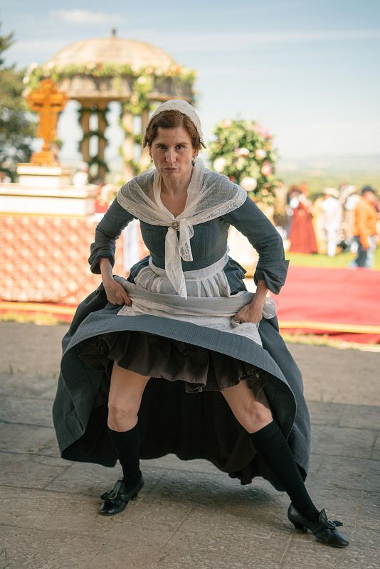 Mujer en vestido gris oscuro y delantal blanco, agachada mostrando sus enaguas, en un jardín con gazebo y personas al fondo.