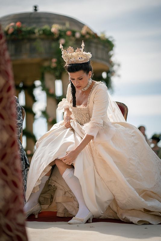 Actriz en traje de época blanco con corona dorada, sentada y arreglándose. Fondo con gazebo decorado y asiento.