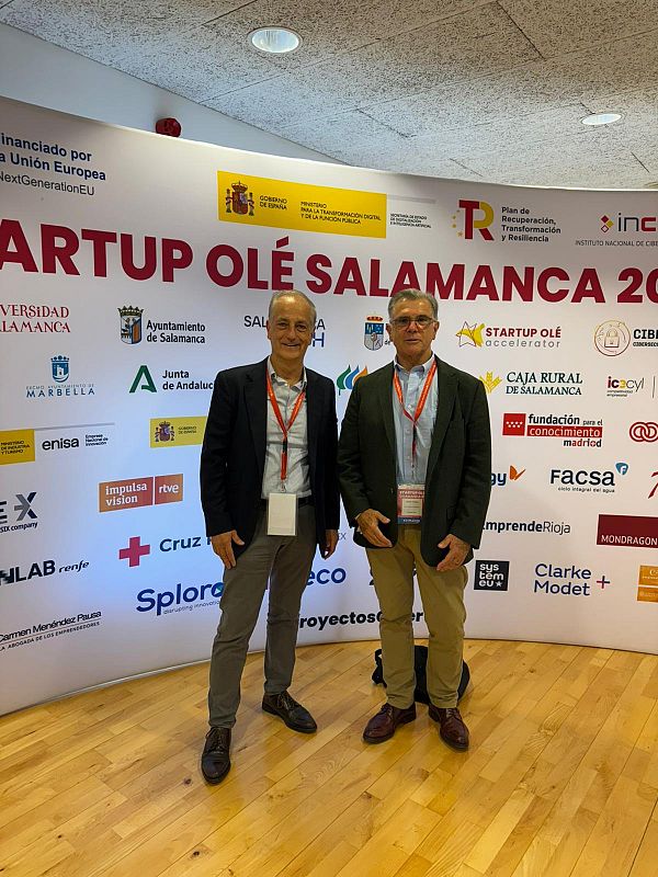 Dos hombres, uno con blazer negro y otro con chaqueta verde oscura, posan frente a un panel con logos y textos. El panel indica financiación europea y el evento "STARTUP OLÉ SALAMANCA 20", con logos de diversas instituciones.