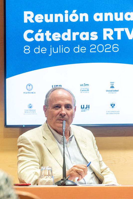 José Manuel Pérez Tornero, con chaqueta beige y camisa azul, ante un micrófono y un vaso de agua. Detrás, pantalla con "Reunión anual Cátedras RTVE" y "8 de julio de 2026", junto a logotipos universitarios.
