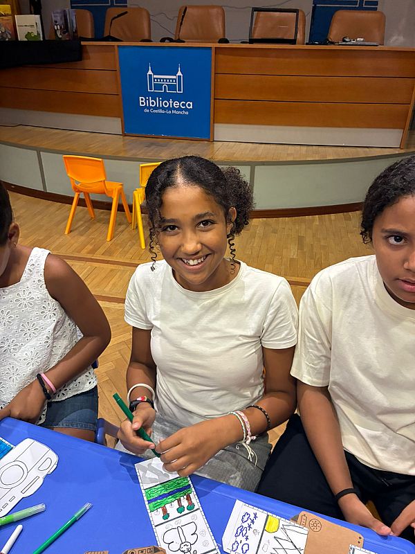 Niñas creando manualidades en la Biblioteca de Castilla-La Mancha. Mansura, con pelo rizado y camiseta blanca, colorea concentrada mientras sonríe a la cámara. Materiales y creaciones de papel sobre mesa azul.
