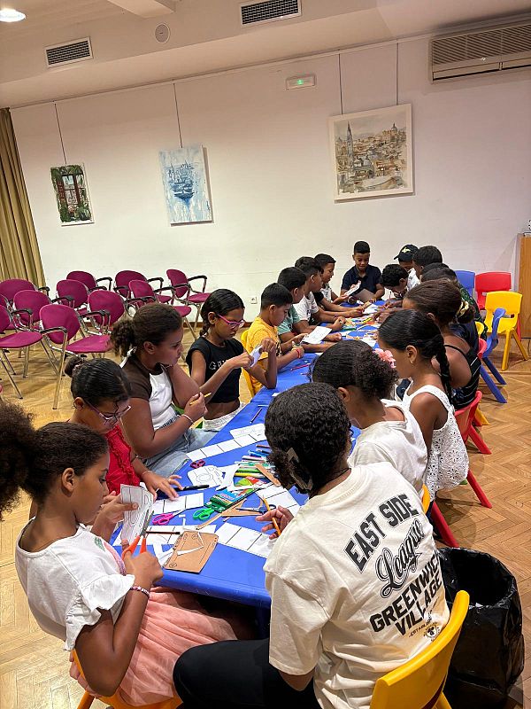 Niños de diversas etnias, entre 6 y 12 años, participan en un taller de manualidades, cortando, pegando y dibujando con materiales variados en una sala amplia y luminosa.