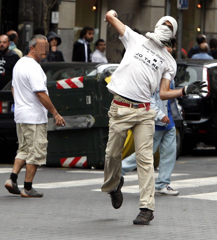 Un encapuchado lanza a una piedra a agentes de la brigada móvil de la Ertzaintza que ha detenido al menos tres personas en los altercados que se han producido este mediodía en el centro de la capital vizcaína.
