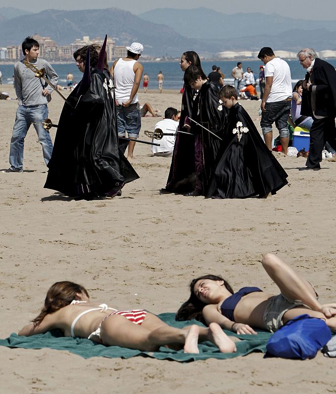 Dos jóvenes toman el sol en la playa valenciana de la Malvarrosa al paso de los integrantes de una cofradía del barrio marítimo del Cabanyal.