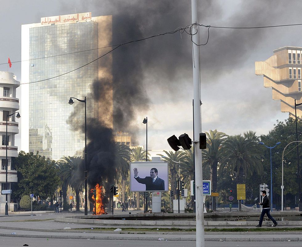 El humo de un incendio causado por unos manifestantes se ve detrás de un cartel del presidente tunecino, Zine el Abidine Ben Alí, en Túnez