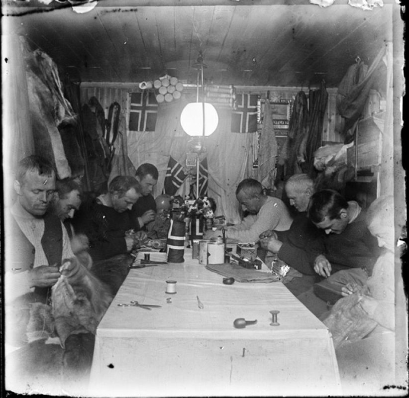 Todos los miembros de la expedición reunidos en torno a una mesa realizando diferentes tareas, principalmente cosiendo sus trajes