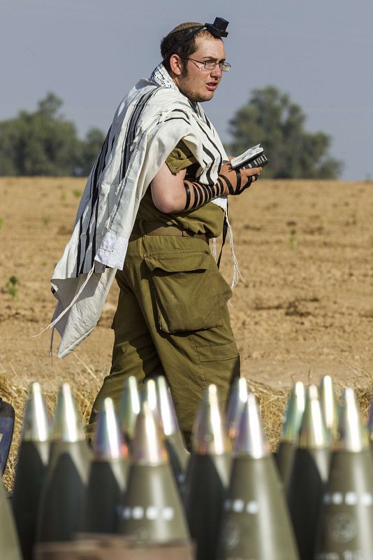 Un soldado israelí portando un 'Talit' (manto religioso) y una filacteria en la frente durante los rezos de la mañana junto a unos proyectiles en la frontera con Gaza