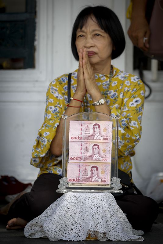 Una tailandesa reza sentada mostrando tres billetes del banco de Tailandia en los que se puede ver la ilmagen del nuevo rey Maha Vajiralongkorn durane la ceremonia de coronación. 