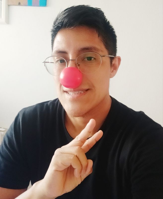 Enrique sonríe y hace el símbolo de la victoria con la mano. Lleva gafas y una nariz roja de payaso. Luce una camiseta negra y el pelo negro y corto.  