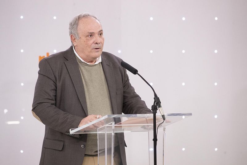 Hombre con americana marrón y jersey beige habla en un atril transparente durante un evento.