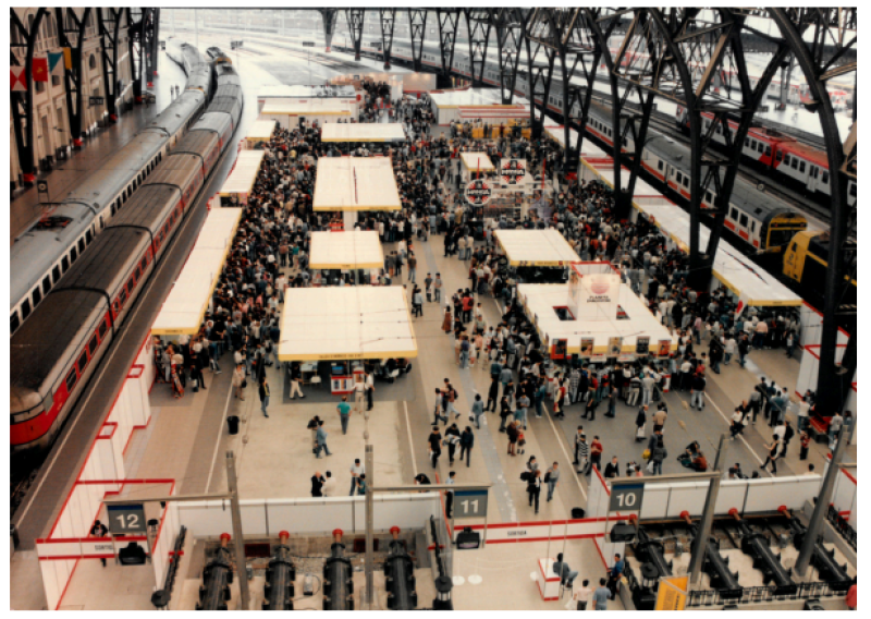 Imagen aérea de un concurrido evento en una estación de tren, con numerosos stands y multitud de personas. Se observan estructuras numeradas (10, 11, 12).
