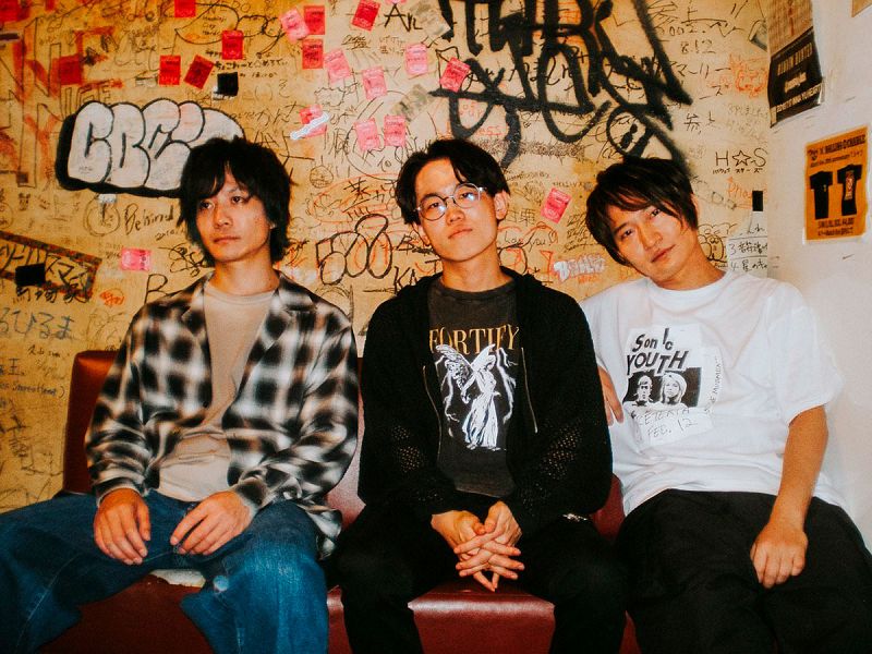 Tres jóvenes sentados, uno con camisa a cuadros, otro con camiseta "FORTIFY" y el último con camiseta Sonic Youth. Ambiente informal con grafitis de fondo.