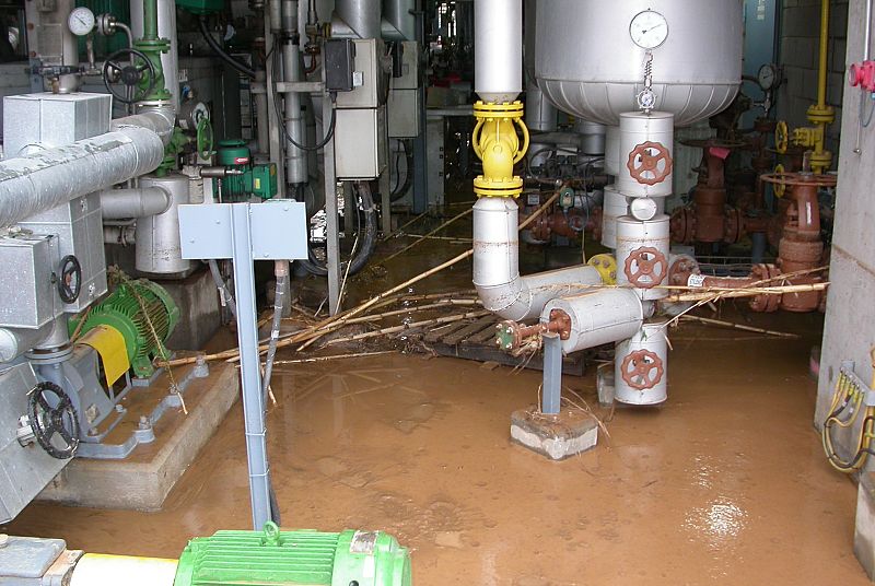 Planta industrial inundada. Agua marrón cubre el suelo, rodeando maquinaria y tuberías.  Restos de vegetación visibles.