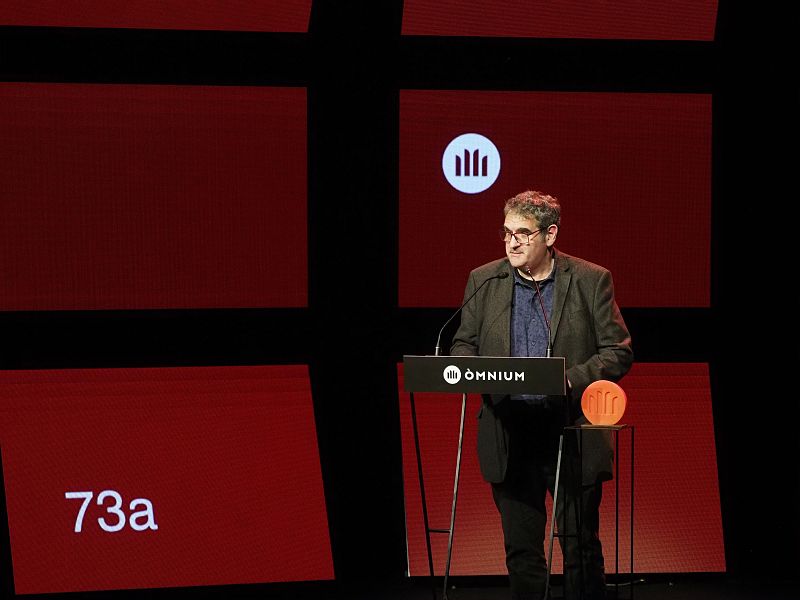 Jordi Puntí, en la 73ª edición del Premi Sant Jordi de novel·la, recibe el premio.  Viste chaqueta oscura y camisa azul marino, y se encuentra de pie junto a un atril con el logo de Òmnium Cultural.