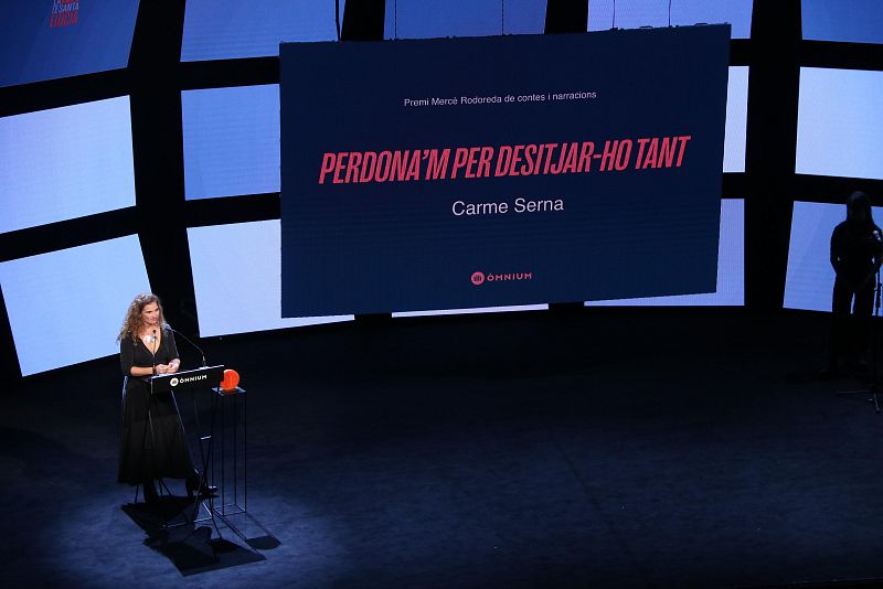 Carme Serna en un escenario con una gran pantalla mostrando el título de su obra y el logo de Òmnium Cultural.  Viste un vestido largo oscuro.
