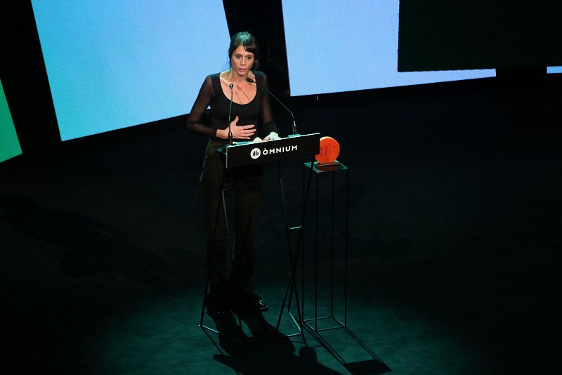 Poeta dando un discurso tras un atril con logo de Òmnium Cultural.  Viste de negro y recibe un premio naranja sobre un pedestal. Iluminación azul y verde en el fondo.
