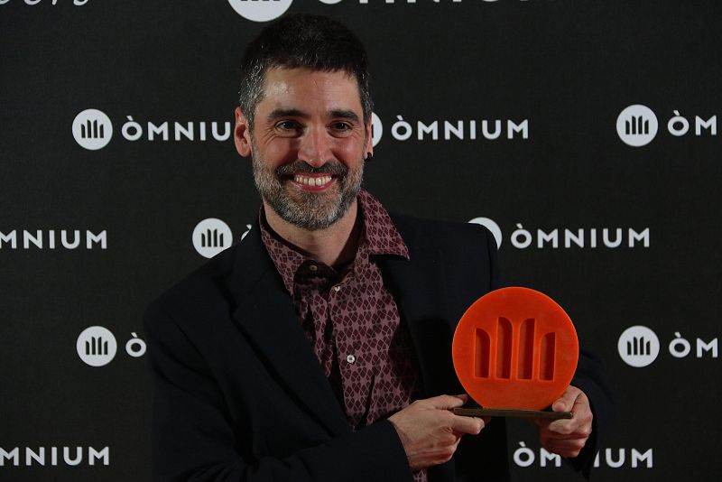 Roc Casagran, con chaqueta oscura y camisa estampada, sonríe al sostener el premio naranja del 65º Premio Sant Jordi de novela. Fondo negro con logo repetido.