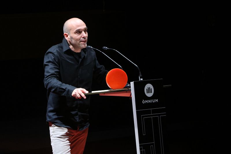 Autor con camisa negra y pantalones claros, de pie ante un atril con micrófono, durante una ceremonia de entrega de premios. El atril muestra el logo de Òmnium Cultural.